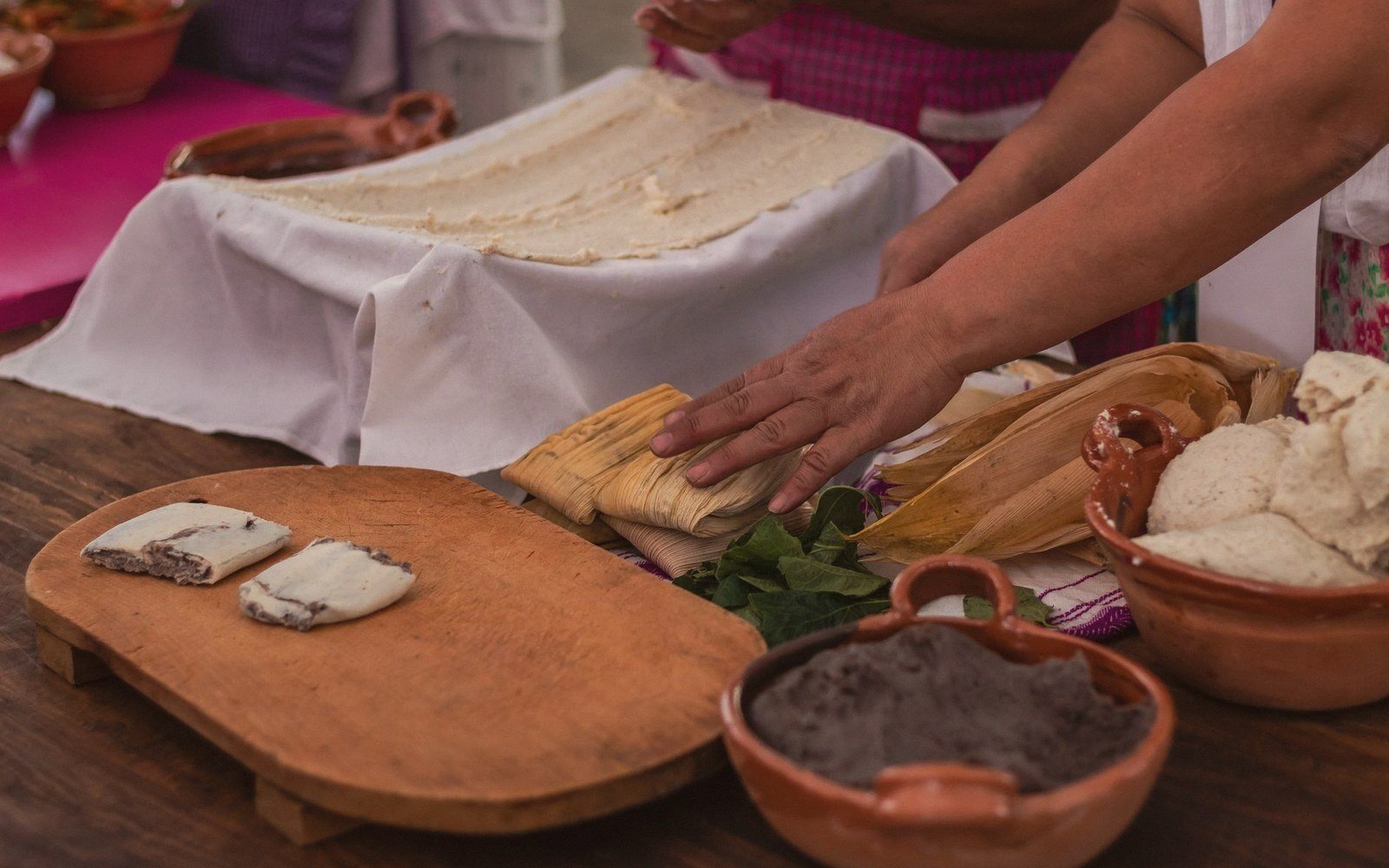 Hands assembling corn husk tamales beside bowls of masa and black bean filling