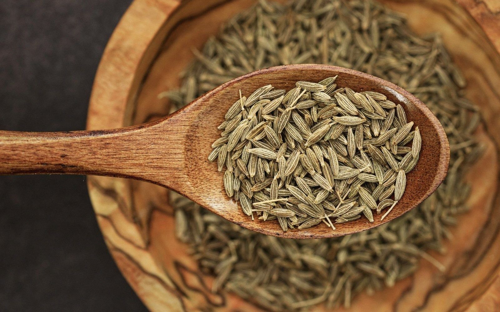 Wooden spoon heaped with cumin seeds resting in a bowl of more seeds
