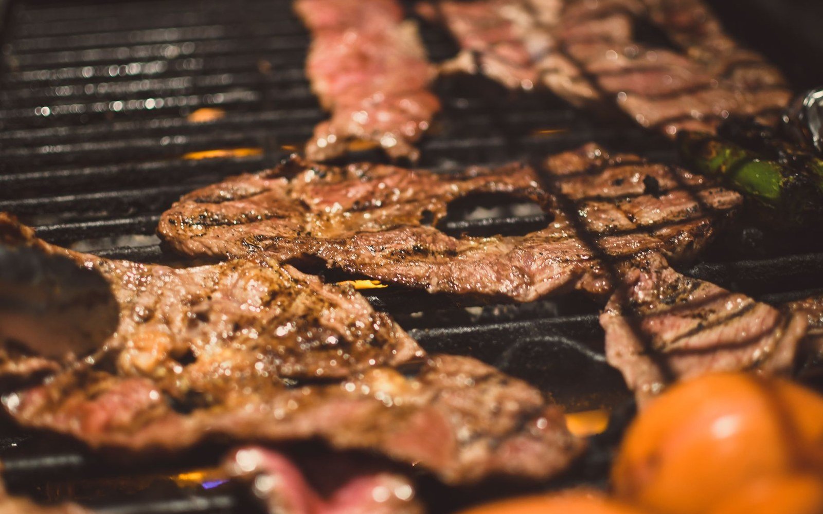 Thin cuts of beef sizzling on a charcoal grill alongside whole tomatoes