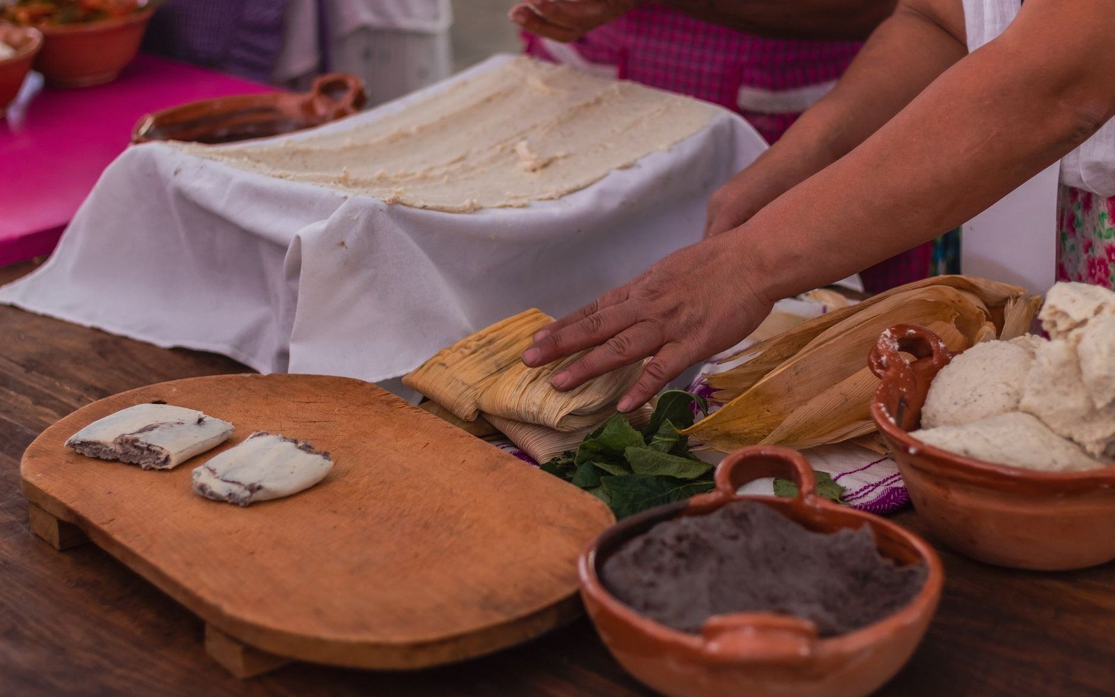 Hands assembling corn husk tamales beside bowls of masa dough and black bean filling