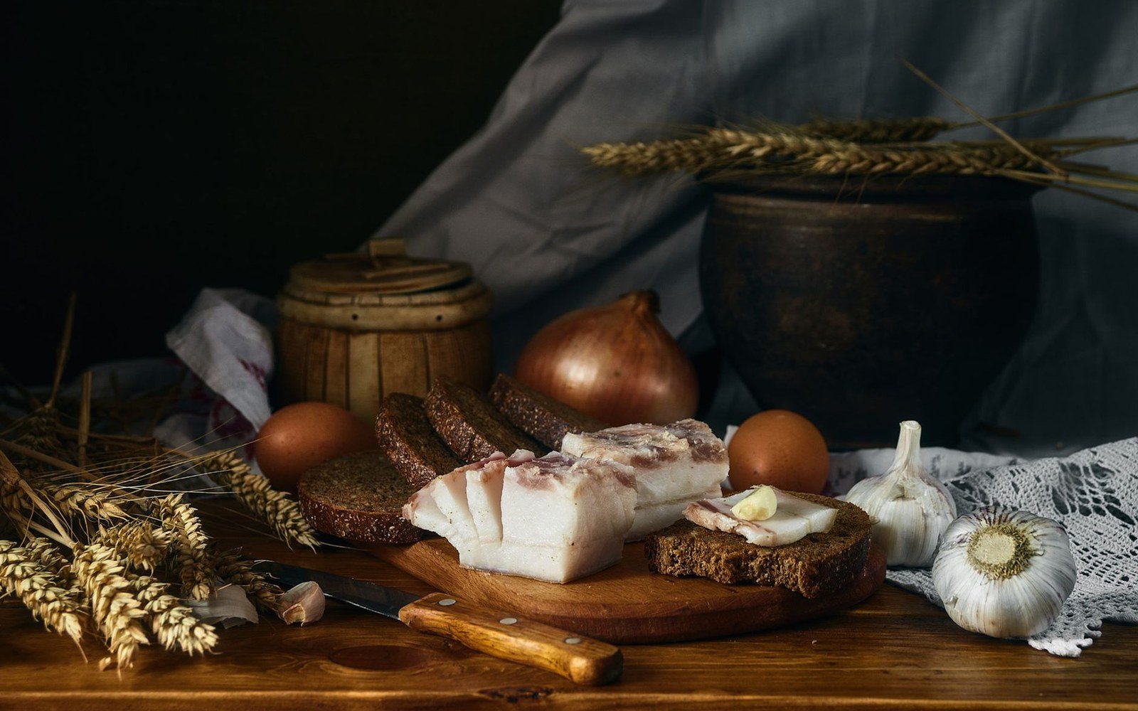 Sliced pork fatback and rye bread on a wooden board with eggs, onion, and garlic