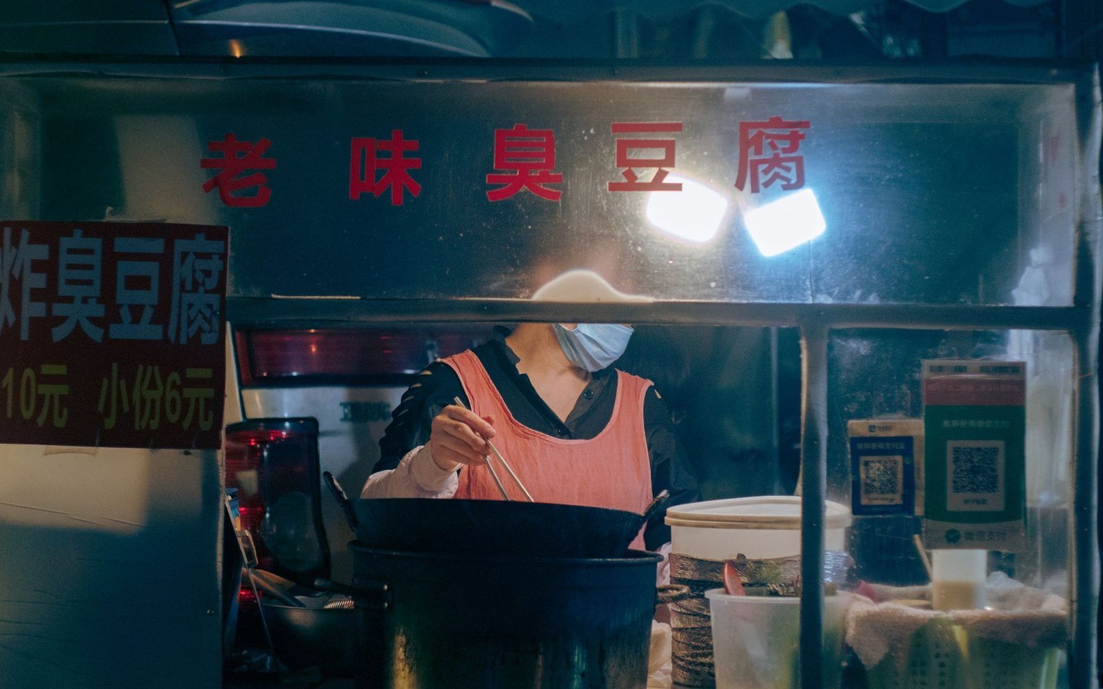 A vendor in a face mask fries stinky tofu at a night market stall