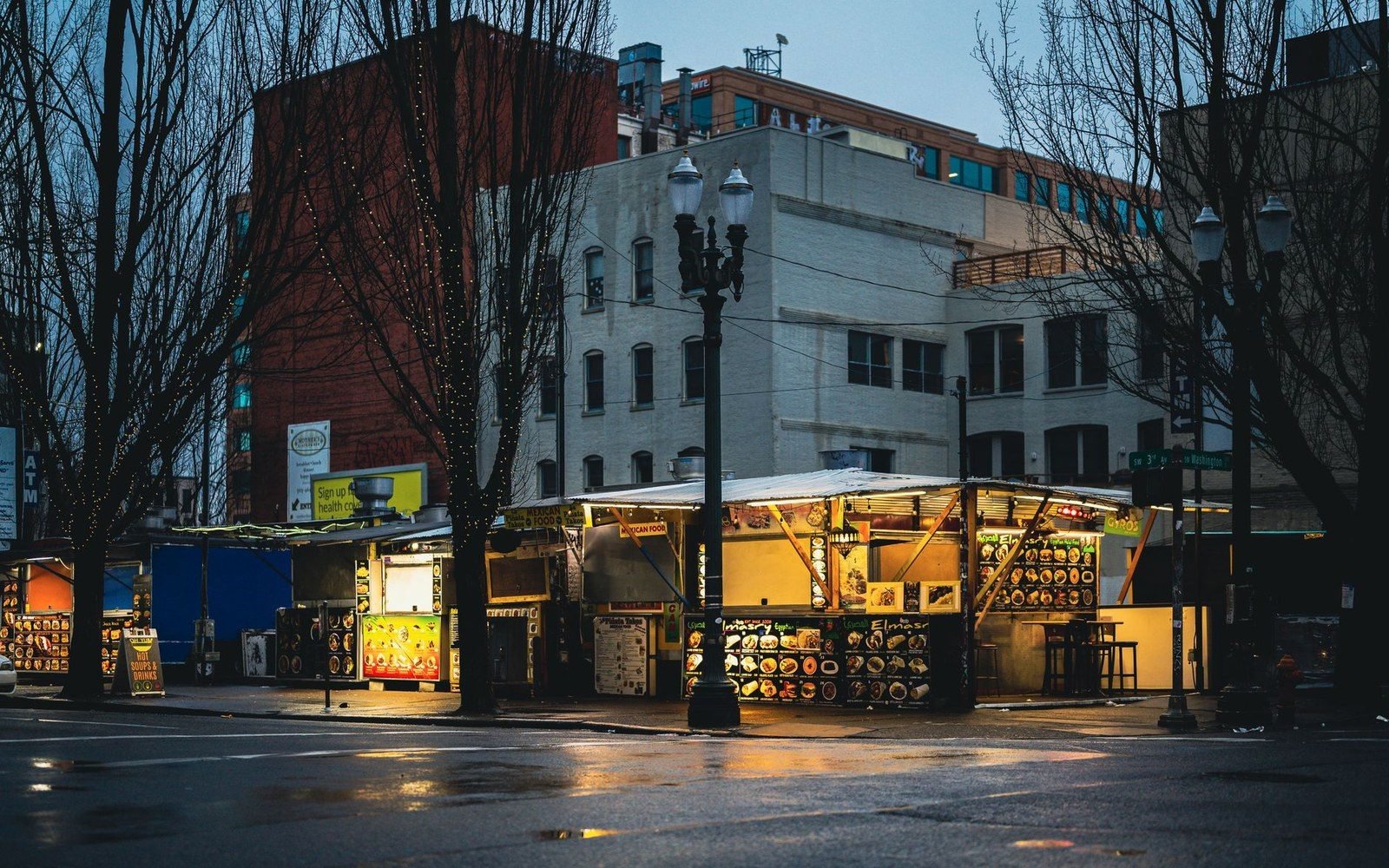 Illuminated food stalls sit on a city corner beneath streetlights at dusk